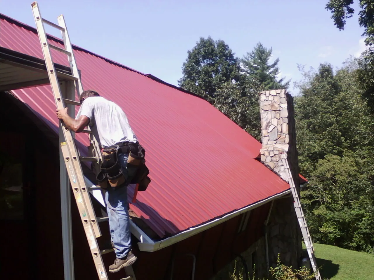 Roofer on a ladder beside a red metal roof and stone chimney — Soffit & Fascia Repair by Roofing Brothers in Beccaria